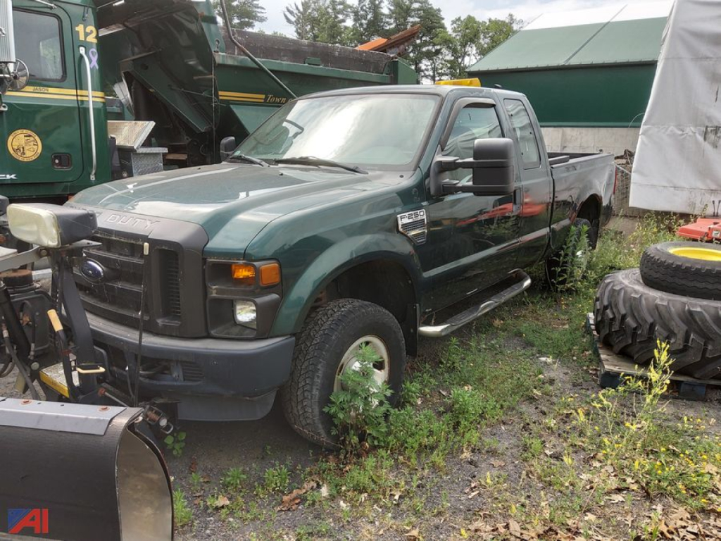 Ford F-250 Super Duty pickup truck with plow attachment in a yard; green exterior with extended cab, large side mirrors, and plow gear visible in front; various tires and equipment around.