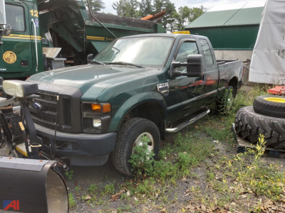 Ford F-250 Super Duty pickup truck with plow attachment in a yard; green exterior with extended cab, large side mirrors, and plow gear visible in front; various tires and equipment around.