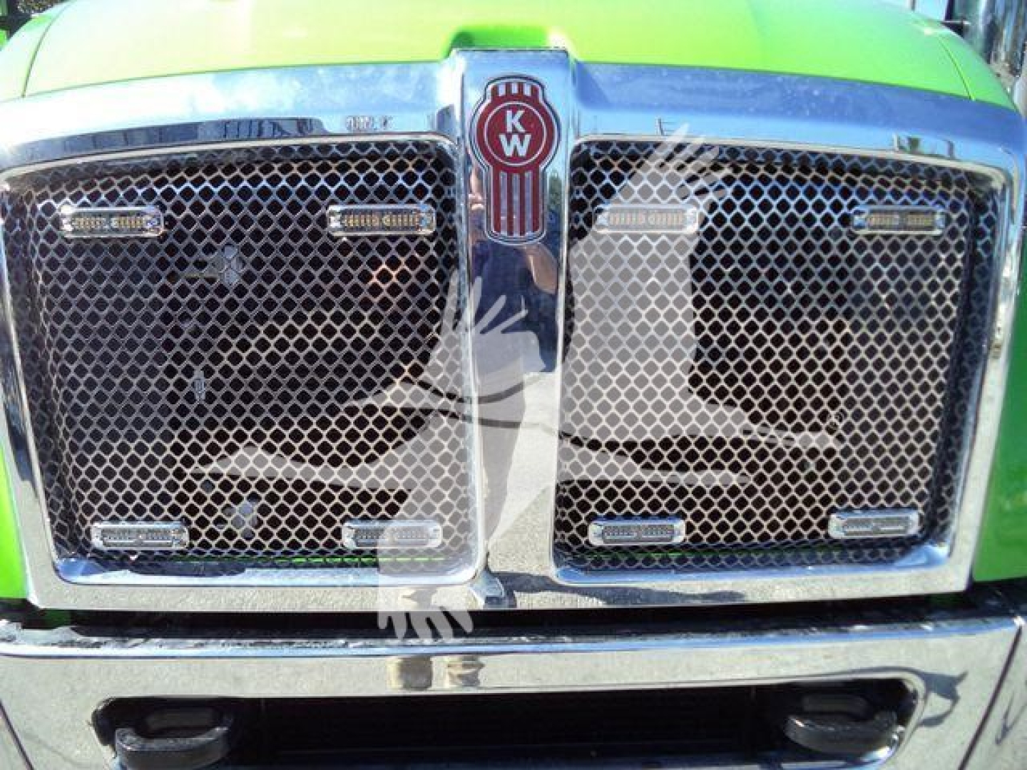 Kenworth T-Series grille close-up showing dual mesh grilles and KW emblem center, bright green paint, chrome trim.