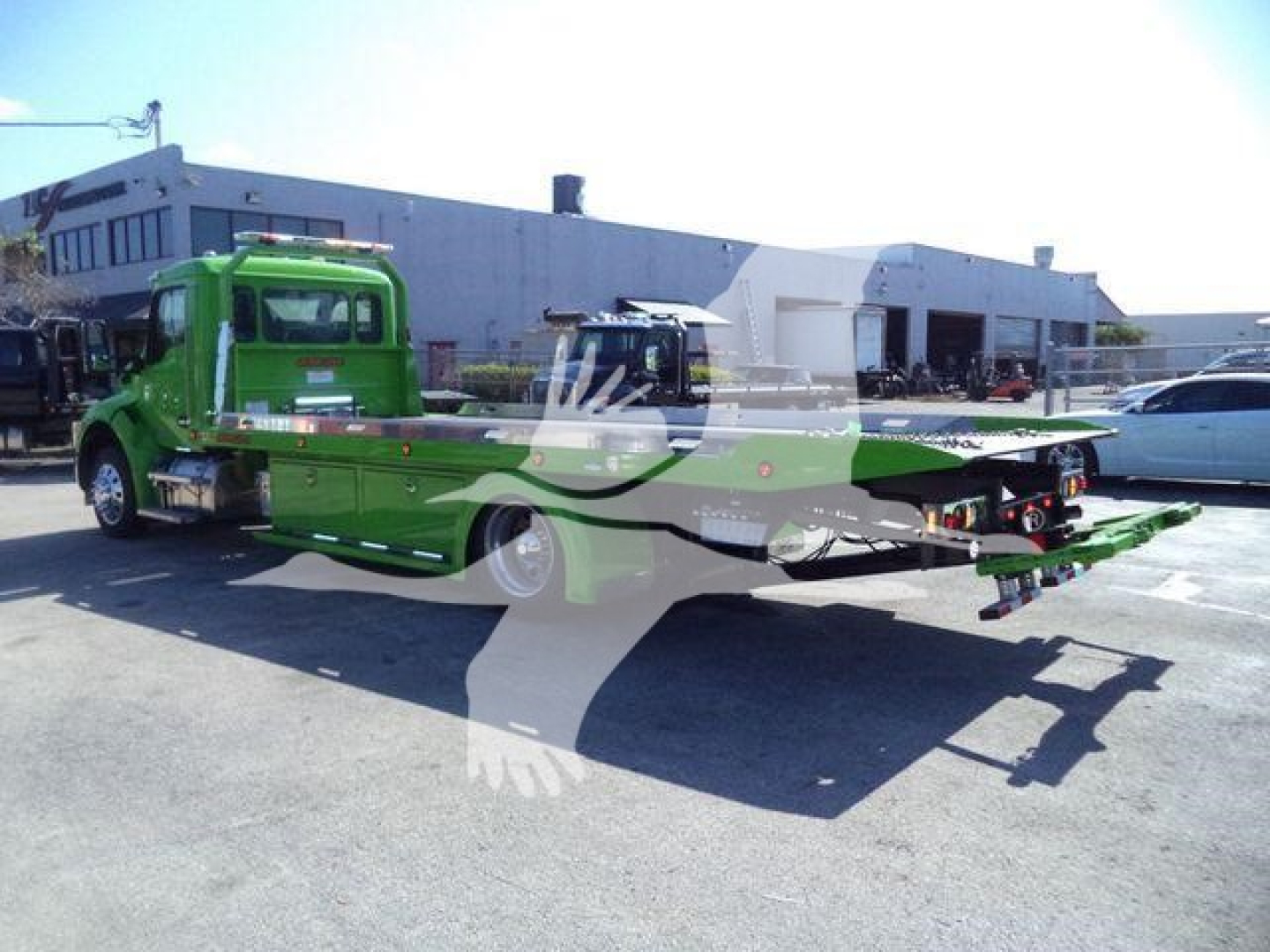 Green Kenworth rollback wrecker truck with flatbed in a lot; visible text on side panels and dealership building in background.