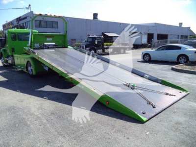 Kenworth T280 rollback truck with a green and silver/metallic ramp bed extended outward, in a dealership lot with other vehicles in background.
