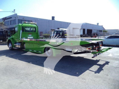 Green Kenworth rollback wrecker truck with flatbed in a lot; visible text on side panels and dealership building in background.