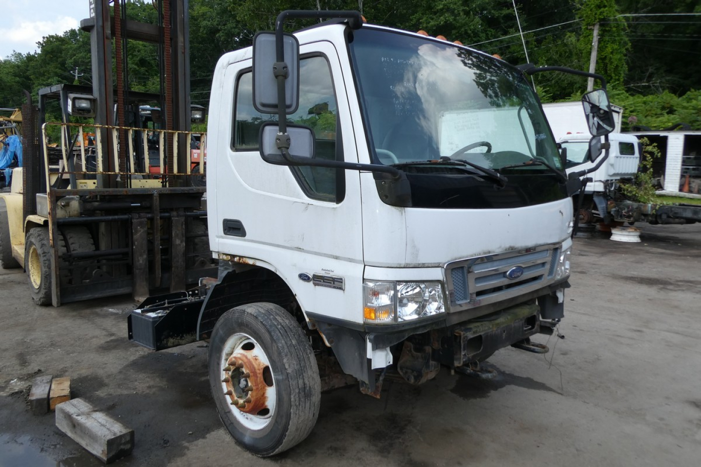 2006 Ford LCF cab and chassis truck front view showing faded white cab, large side mirrors, and exposed front bumper; visible Ford emblem on grille and headlamps; white cab with black trim; background includes other equipment.