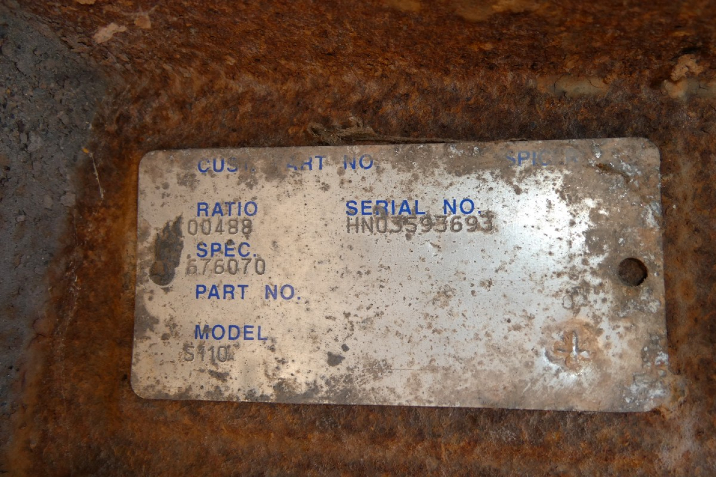 Plate with worn blue text on white background containing fields like SERIAL NO, SPEC, MODEL, PART NO, RAT... on a rusted surface.