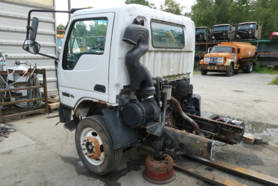 Ford LCF cab and chassis truck with white cab, exposed engine area, and wheel removed; shows side view with nearby equipment and other trucks in background.