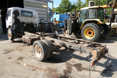 Truck chassis frame with rear wheels and exposed driveline in a yard, surrounded by construction and industrial vehicles.