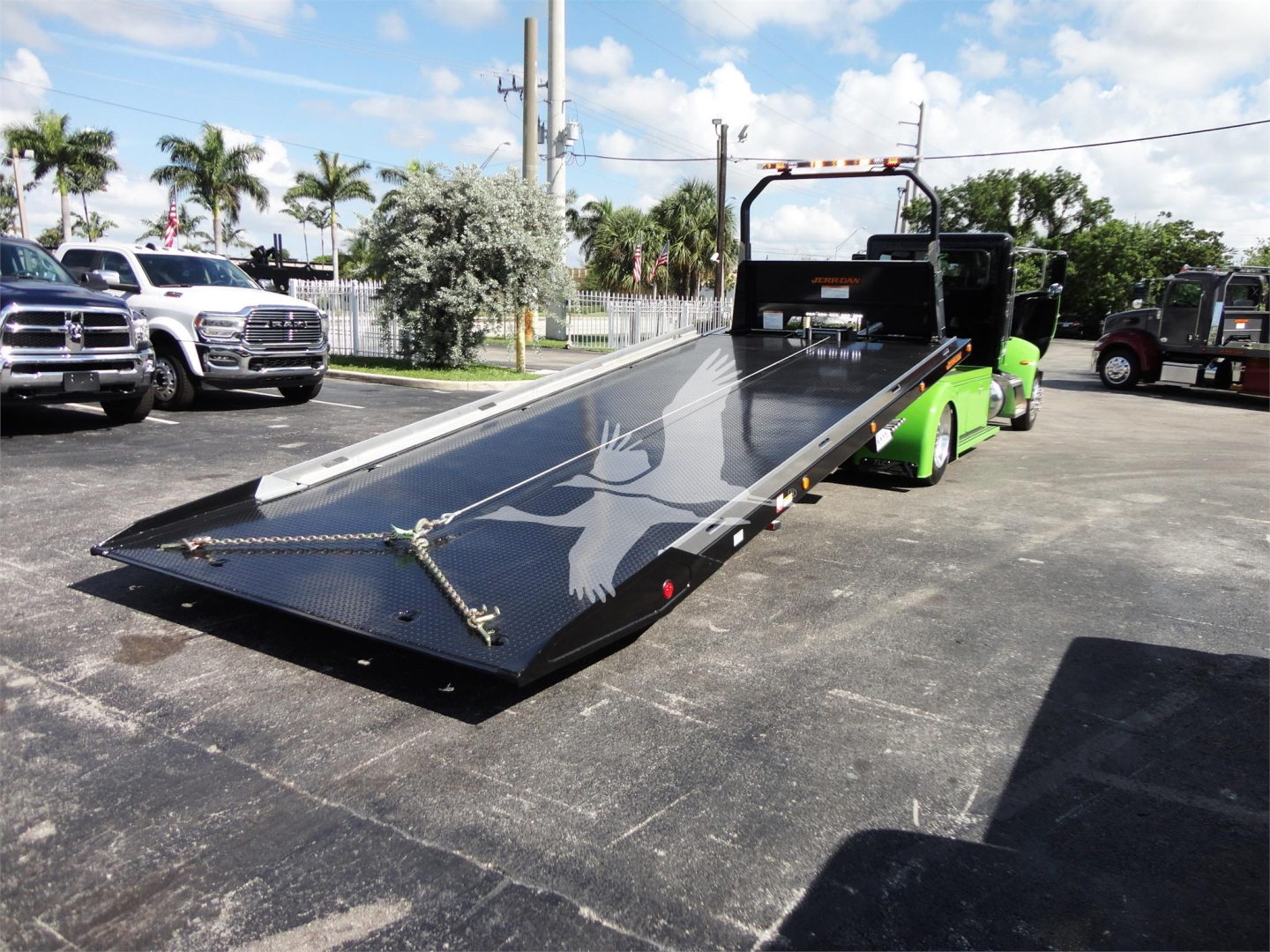 Truck with a flatbed rollback trailer on a parking lot, showing a black metal ramp and green tow vehicle.