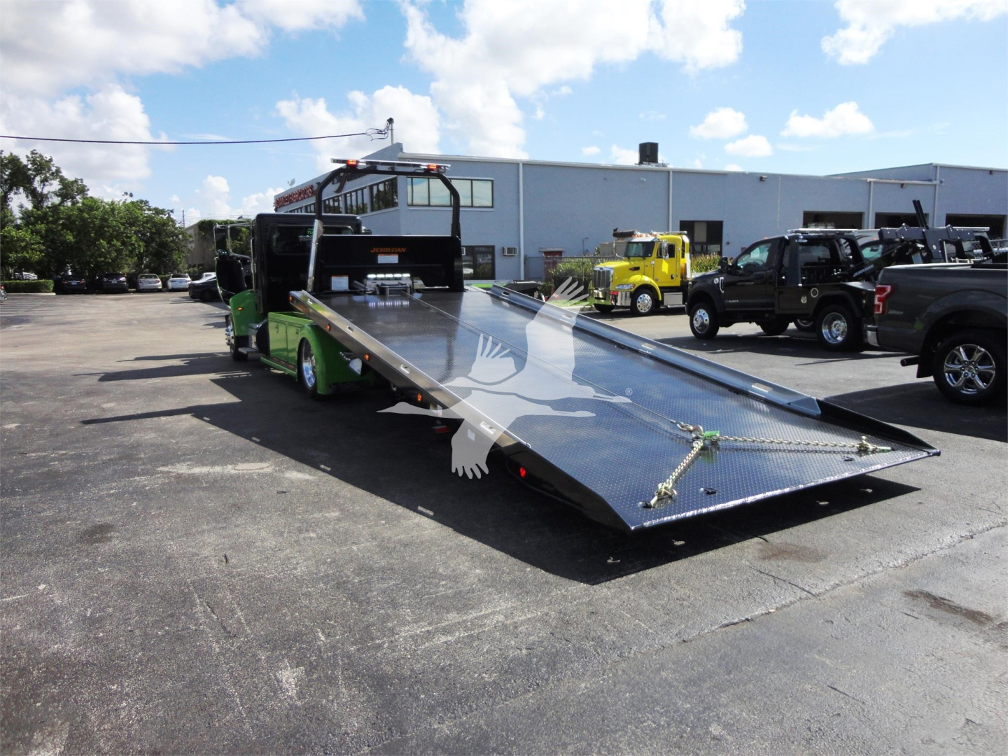 Tow truck with a long metal flatbed ramp in a parking lot, Peterbilt 337 chassis in background, several other trucks nearby.