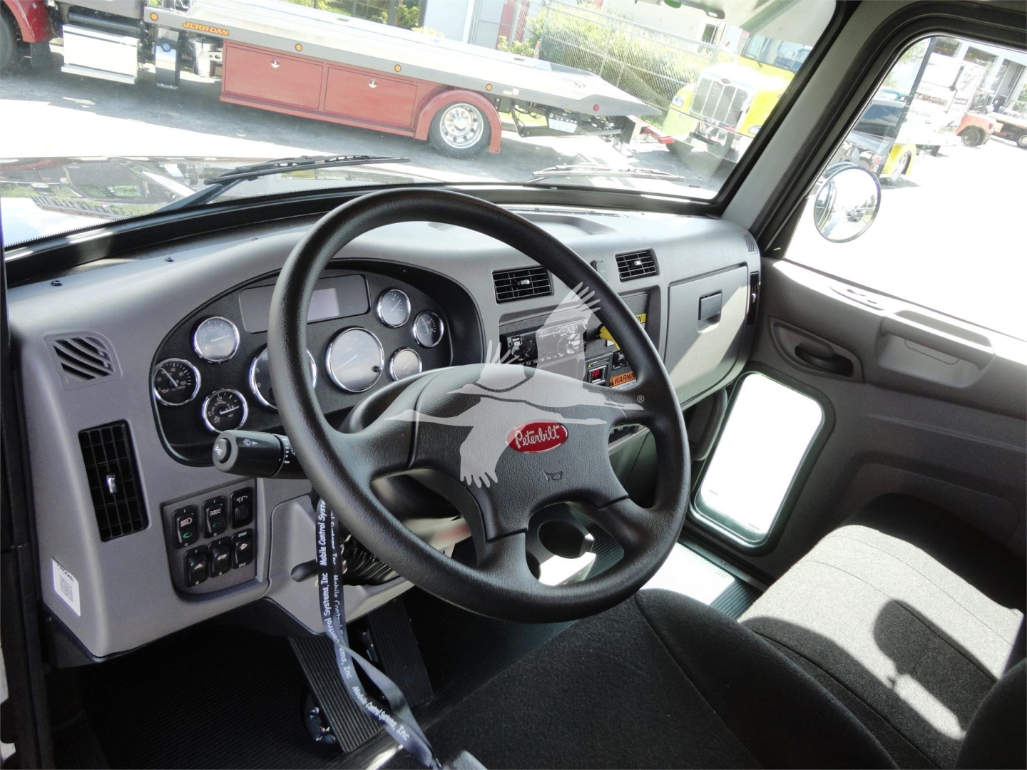 Peterbilt truck cockpit interior with steering wheel and instrument cluster; dashboard controls visible; side window and rearview mirror showing parking lot.