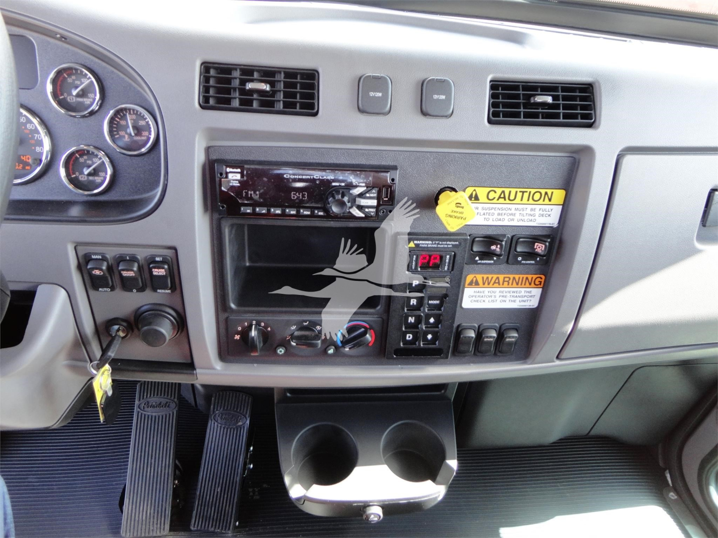 Dashboard and center console of a Peterbilt 337 cab showing radio head unit, climate controls, warning stickers, cup holders, and pedal area.