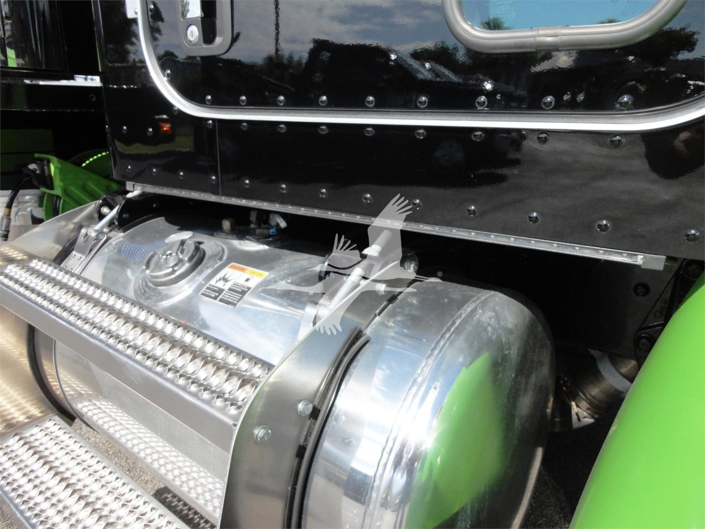 Peterbilt 337 truck engine bay showing a polished cylindrical fuel tank with chrome cap and diamond-plate step; side of cab is black with riveted trim and green frame visible.
