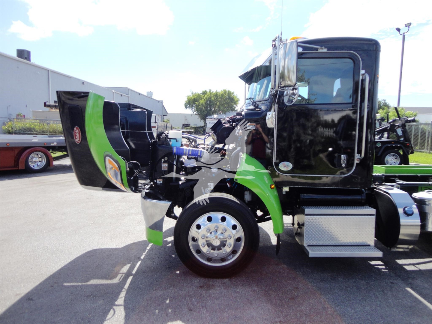 Truck cab side view with engine hood open showing green and black Peterbilt cab, chrome wheel, aluminum steps, and front fender area.
