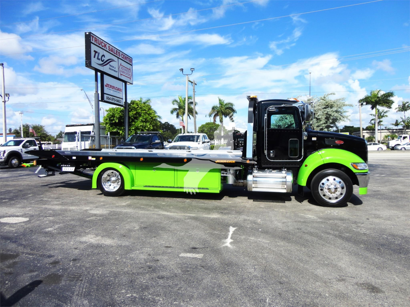 Flatbed tow truck in lime green and black, side view. Visible text on signage in background reads TLC or similar dealership signs. No legible model or serial numbers on truck visible in image.
