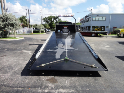 Flatbed tow truck bed with ramp at rear, Peterbilt 337 not visible in cab, watermark of bird silhouette on ramp. Scene shows dealership lot with other trucks and buildings.