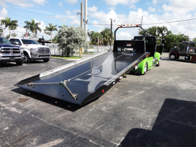 Truck with a flatbed rollback trailer on a parking lot, showing a black metal ramp and green tow vehicle.