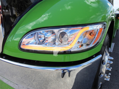 Green Peterbilt truck headlight area with chrome bumper and wheel, close-up front-left view.