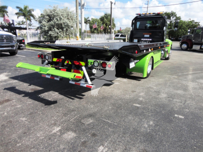 Green Peterbilt 337 rollback tow truck viewed from rear-left with hydraulic flatbed and rear taillights visible.