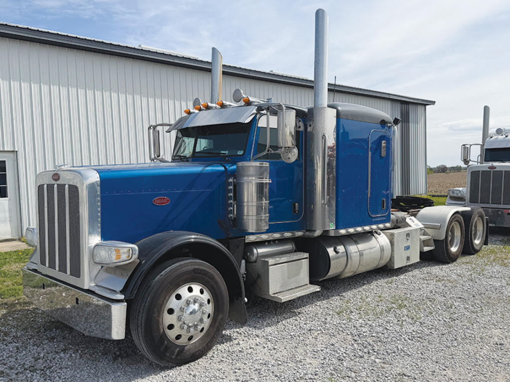 Peterbilt 389 conventional sleeper truck in blue, front grille, tall vertical exhaust stacks, chrome fuel tanks, side steps, and dual rear axles parked on gravel.
