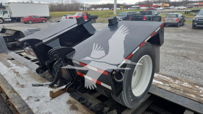 Trailer showing dual-axle lowboy with black metal frame and white wheel, top deck with textured surface, hydraulic ramps, and safety chains; some equipment visible near the hitch.