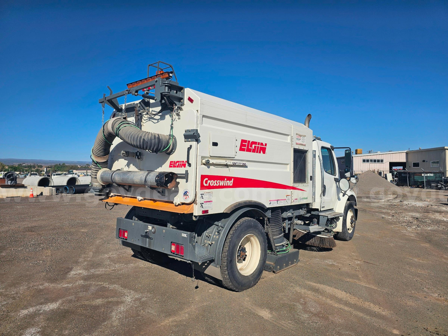 Back view of a white Freightliner M2 106 sweeper truck with Elgin Crosswind branding and exposed rear machinery in a dirt lot.