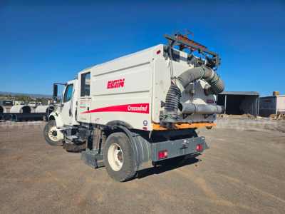 Back-right view of a Freightliner M2 106 sweeper truck with Elgin crosswind system, white cab and white dump body, large exhaust hose, and rear hose assembly.