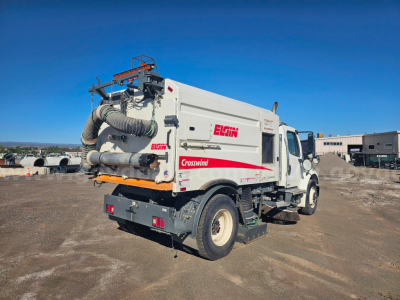 Back view of a white Freightliner M2 106 sweeper truck with Elgin Crosswind branding and exposed rear machinery in a dirt lot.