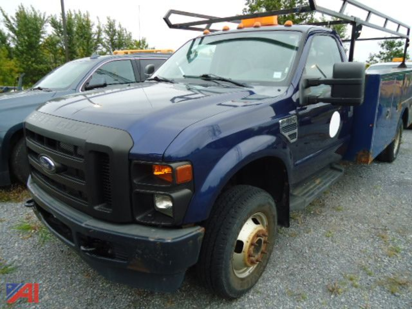Ford F-350 Super Duty truck with utility bed and dual rear wheels, blue cab, black grille, front bumper, amber lights on roof, and rack in background.
