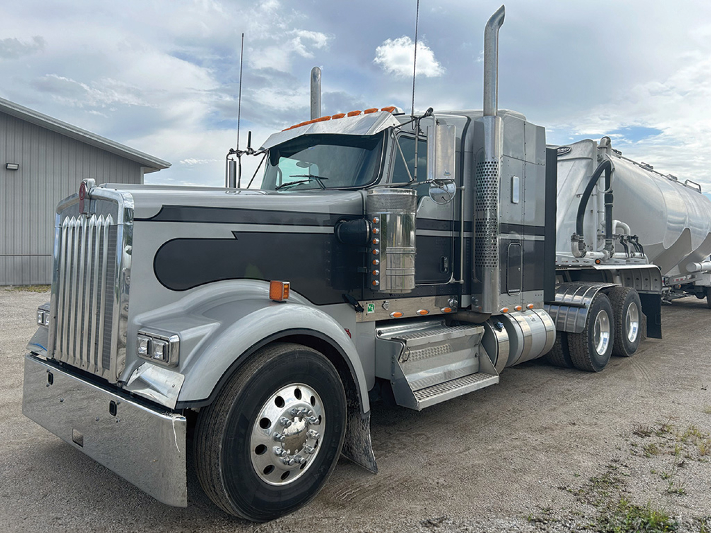 Kenworth W-900L AeroCab conventional sleeper truck in metallic gray, chrome accents, large grille, dual exhaust stacks, and a tanker trailer attached.