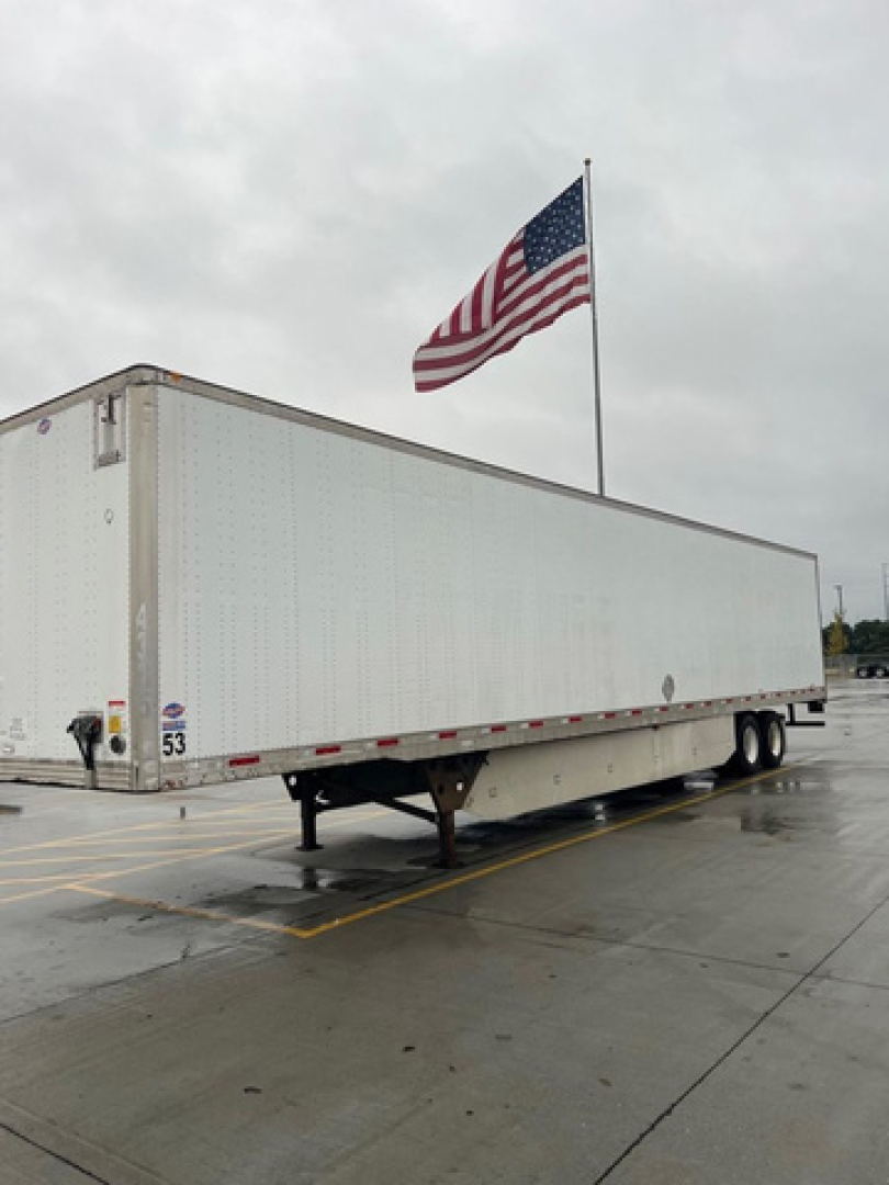 Utility van trailer in a parking lot with American flag flying on a pole.
