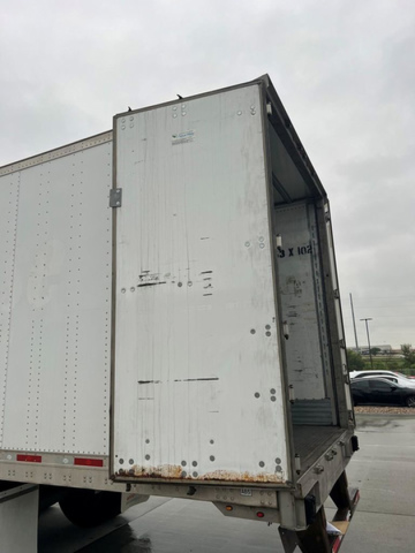 Rear of a utility trailer with open rear doors showing a metal box and interior. Visible text includes small label on door and scuffs.