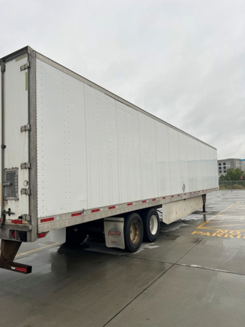 Trailer side view showing a white utility van trailer with dual rear wheels and reflective tape along the lower edge.