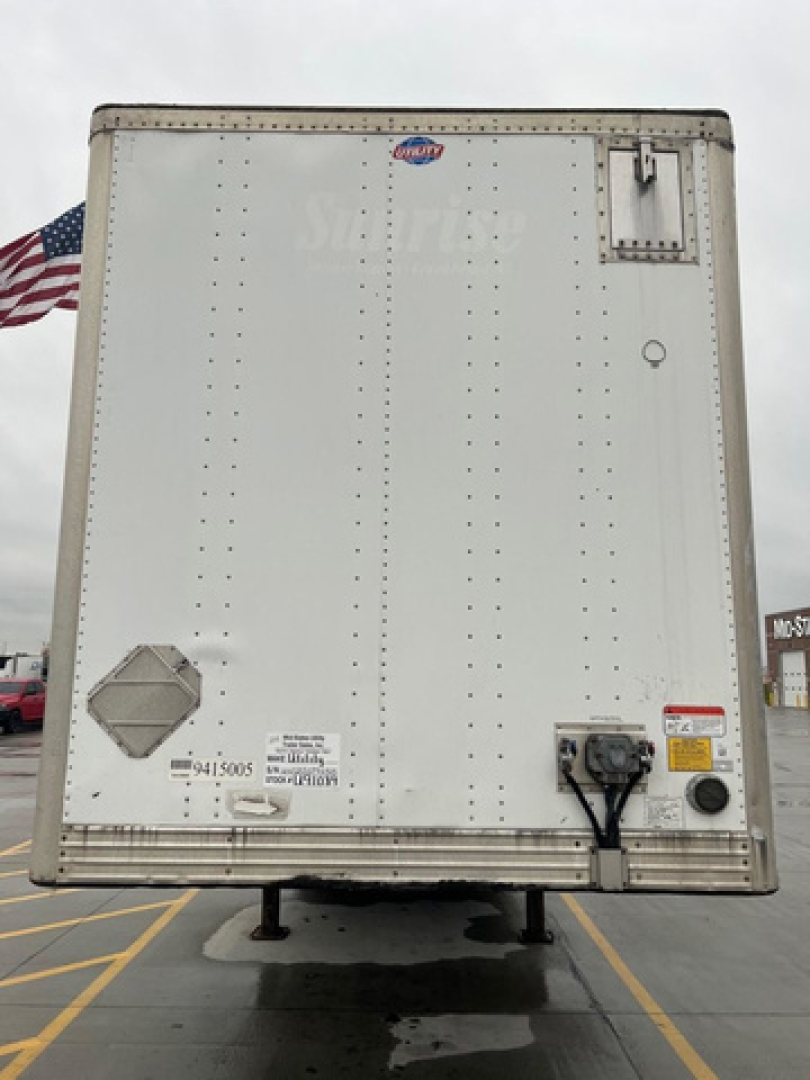 Rear view of a white trailer with riveted panels, small door latch, and electrical connection; USA flag visible on left side.