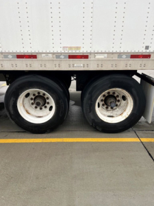 Trailer axle area showing dual rear wheels and lower side of a white utility trailer with reflective tape and wheel rims.