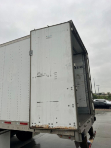 Rear of a utility trailer with open rear doors showing a metal box and interior. Visible text includes small label on door and scuffs.