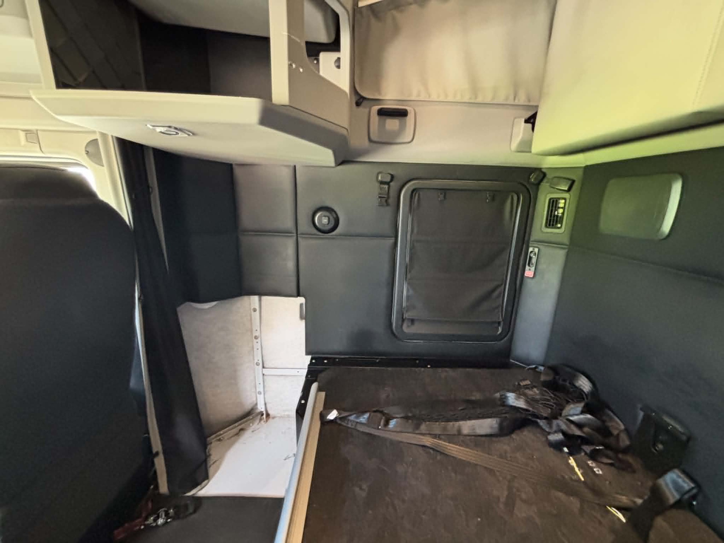Interior cab area of a Kenworth T680 sleeper truck showing a rear cab entry/exit door, overhead storage, and a raised bunk area with seat belts and trim panels.