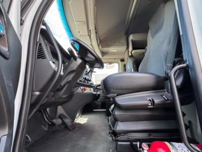 Kenworth truck cab interior showing steering wheel, dashboard, and driver seat with center stack; gray fabric seats, pedals, and entry area visible.