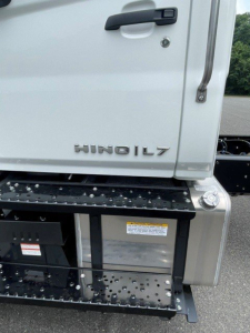 Cab chassis rear-left view showing Hino L7 badge on tailgate and fuel/air tank area with metallic steps and warning label.