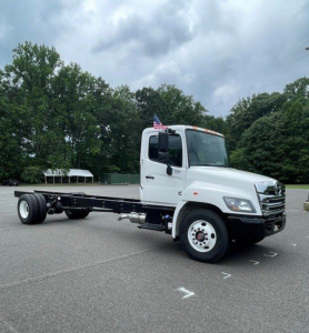 Hino L7 cab chassis truck showing front and side view with white cab and black chassis, no body on bed, US flag on hood area.