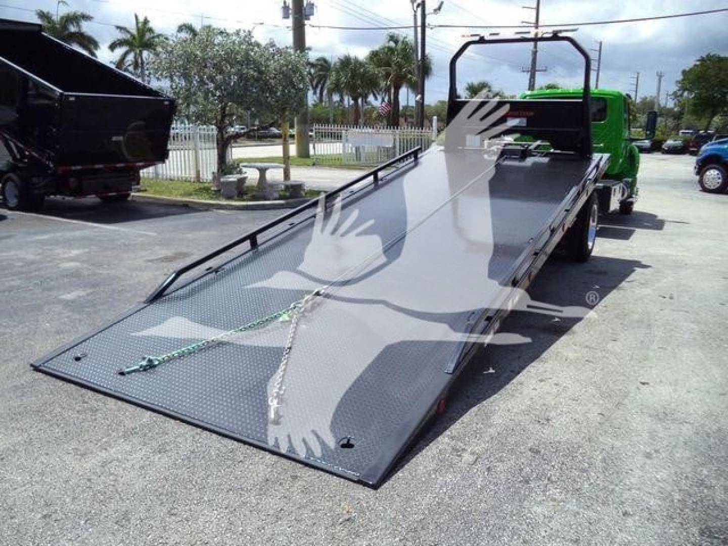 Flatbed tow truck in service bay showing a long black ramp bed with textured surface and green truck cab in background.