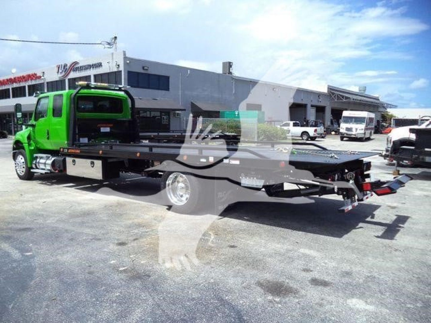 Green tow truck with rollback bed in a dealership lot, International MV model visible with flatbed and loading equipment.