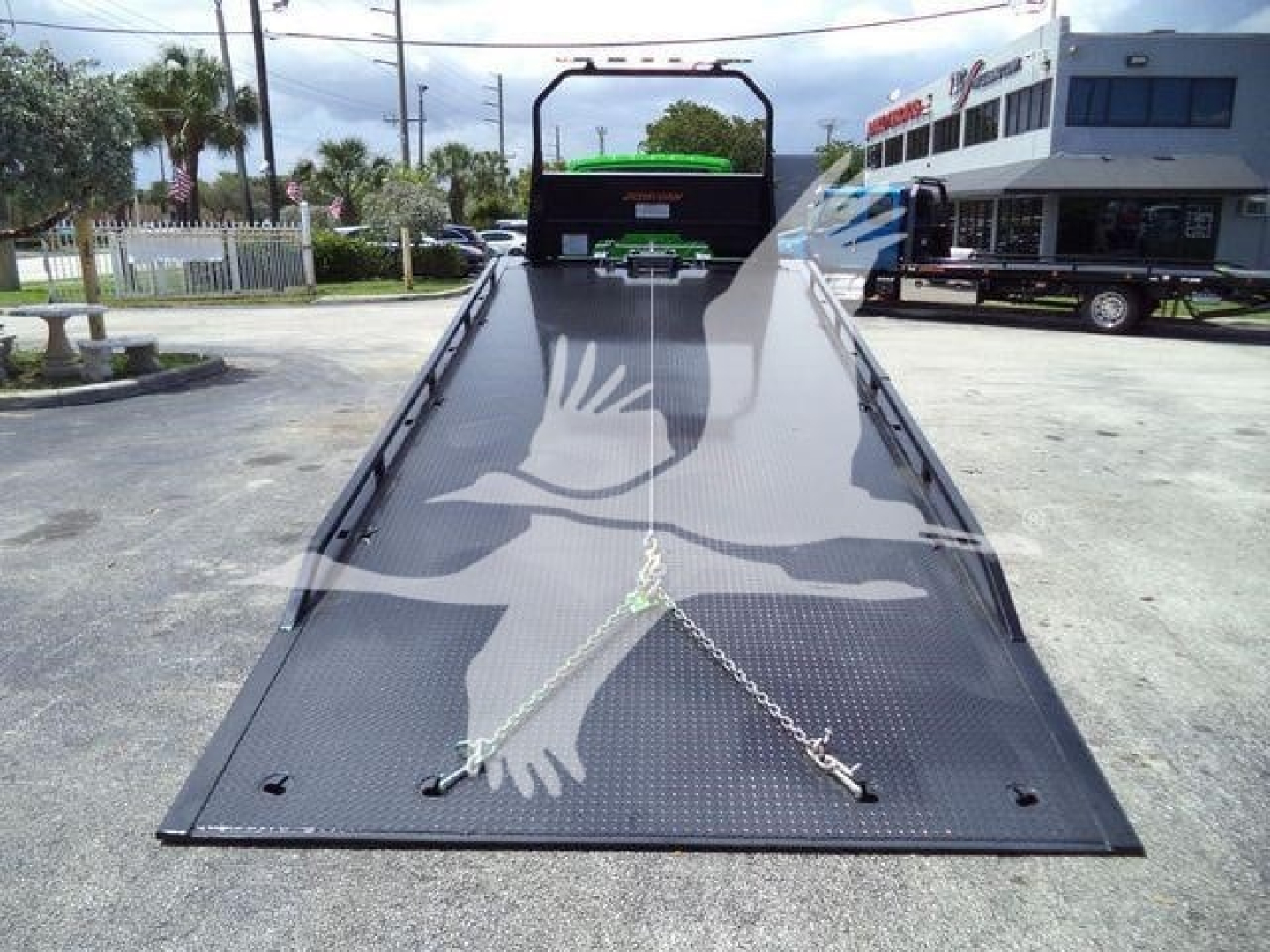 Wide view of a black metal tow rollback bed with chain anchors on the ramp, perspective from the tip of the ramp toward the cab, no text clearly visible on the ramp.