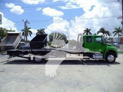 Flatbed rollback tow truck with green cab and black bed, rear loading ramps and a dropped chassis, side view in a dealership lot with palm trees behind