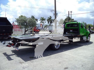 Flatbed tow truck with green cab in a parking lot. Details visible: No legible labels or stickers on the flatbed; small text on tail area not readable.