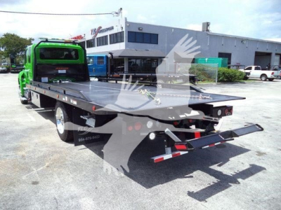 Green International MV rollback tow truck with flatbed in rear, viewed from left-back angle; low rear ramps extended; dealership building in background.