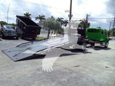 Truck bed with a lowered flatbed tow ramp, green tow truck on right, black dump truck in background, outdoor lot.
