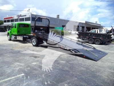 Truck with a green cab and a long black tow ramp attached to a rollback bed, in a dealership lot.