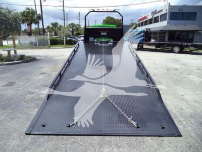 Wide view of a black metal tow rollback bed with chain anchors on the ramp, perspective from the tip of the ramp toward the cab, no text clearly visible on the ramp.