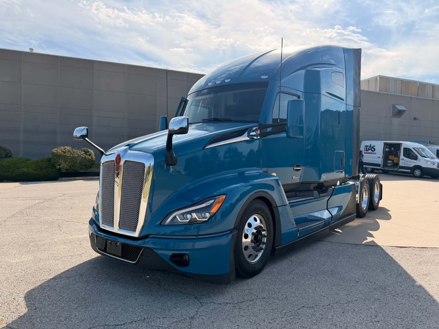 Kenworth T680 sleeper cab in blue, front-left view with grille, headlights, and side mirror visible.