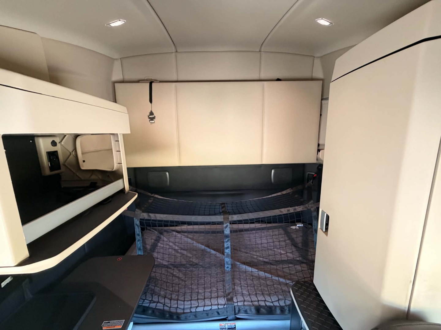Kenworth T680 sleeper cab interior showing bunk area and cargo net behind; clean light-colored panels and overhead storage.