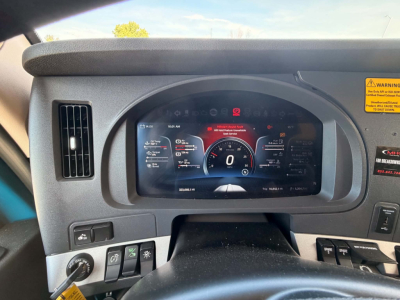 Kenworth T680 dashboard view with digital instrument cluster showing 0 speed, steering wheel in foreground, various controls to left, warning sticker on right.